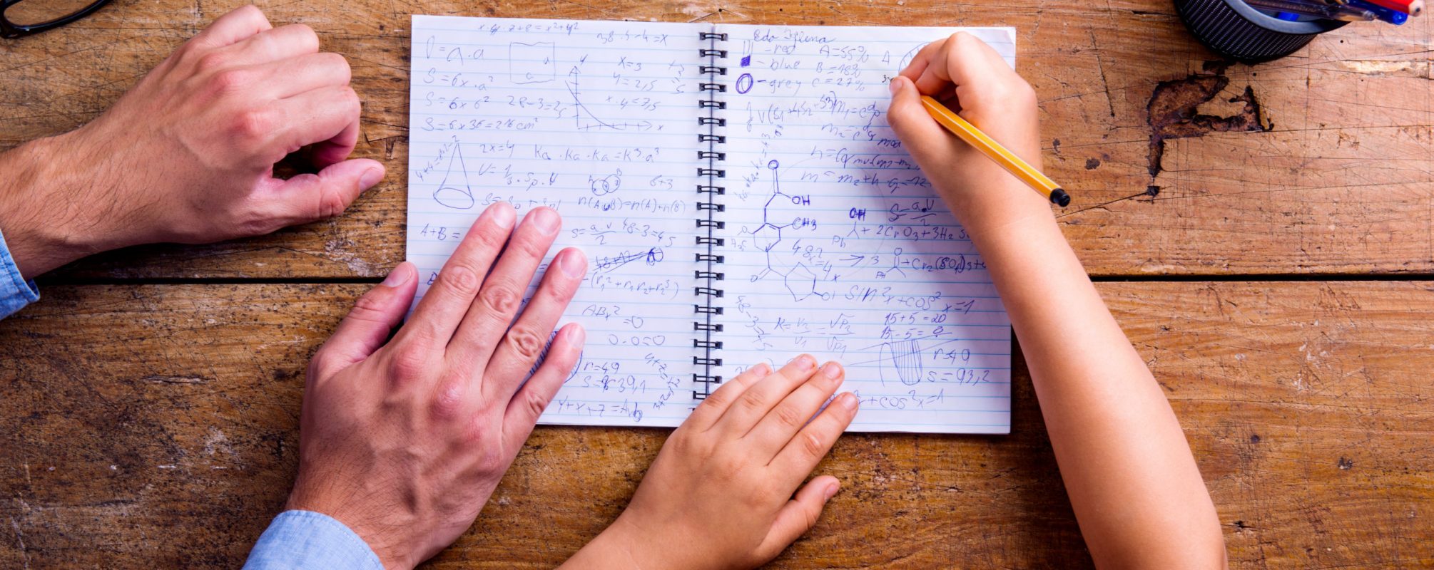 Unrecognizable father and son studying together, writing into a notebook. Studio shot on wooden background. Flat lay.